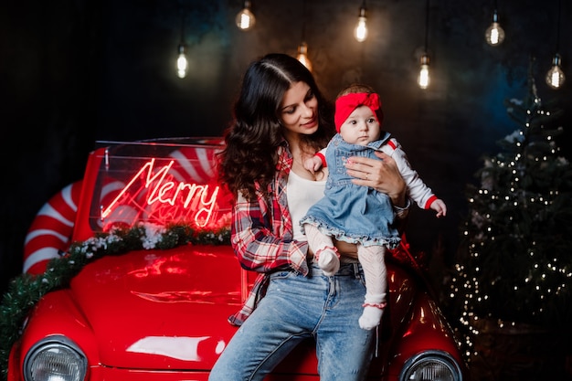 Mulher feliz com uma menina com um laço vermelho na cabeça está sentada e se divertindo em um carro retrô com decoração de Natal