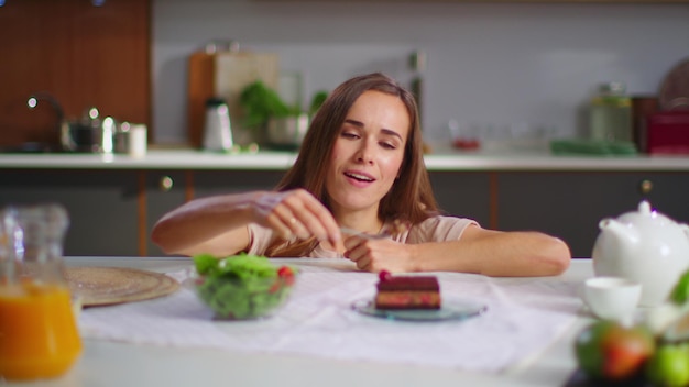 Mulher feliz comendo bolo em vez de salada na cozinha Senhora desfrutando de bolo na mesa