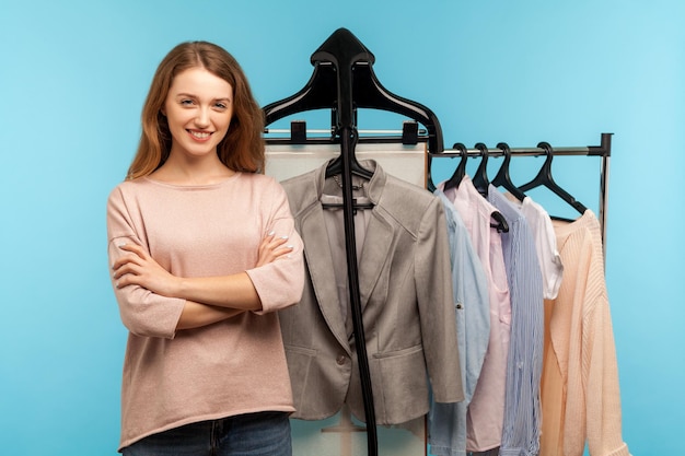 Mulher feliz confiante, estilista de moda profissional especialista em pé perto de roupas penduradas na prateleira na loja de grife, vendendo roupas da moda na loja boutique. tiro de estúdio interno isolado em fundo azul