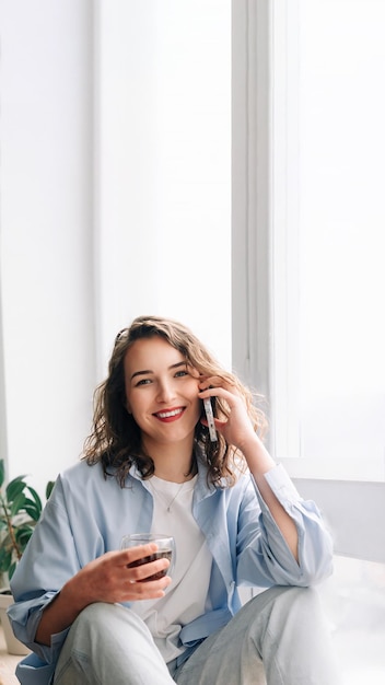 Mulher feliz conversando ao telefone com a irmã ou amiga durante o chá na janela da cozinha agradável