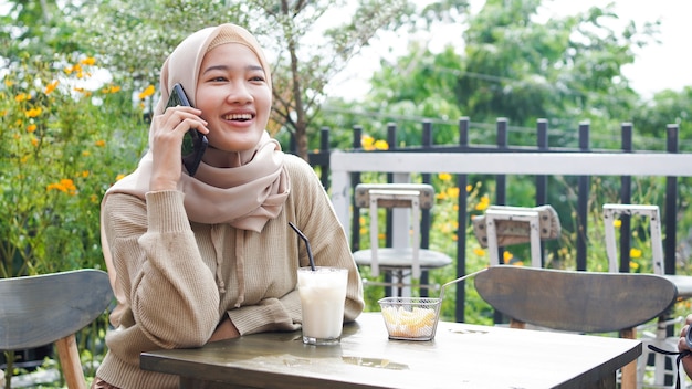 Mulher feliz da ásia hijab usando telefone inteligente e ligando, sentada em um café