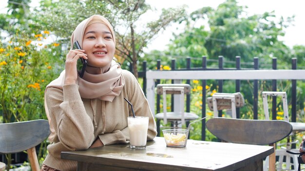 Mulher feliz da Ásia hijab usando telefone inteligente e ligando, sentada em um café