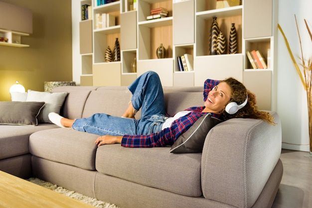 Mulher feliz de camisa xadrez e jeans relaxando no sofá da sala de estar e ouvindo música em fones de ouvido brancos