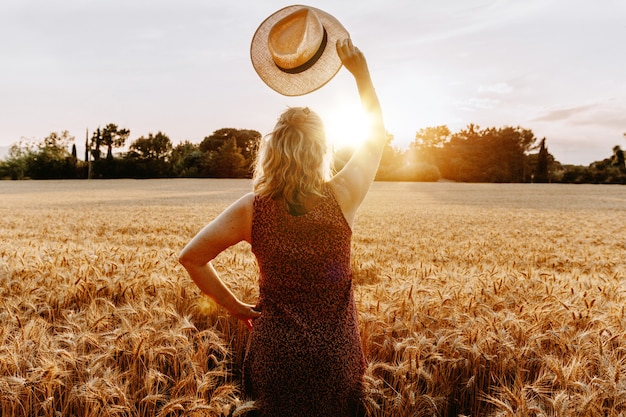 Mulher feliz de meia-idade ou sênior se sentindo livre com um vestido de braços abertos e chapéu dentro de um campo de trigo na temporada de verão