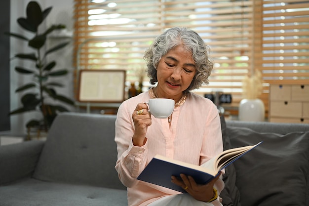 Mulher feliz de meia-idade relaxando no sofá e lendo um livro interessante desfrutando do tempo de lazer de fim de semana em casa