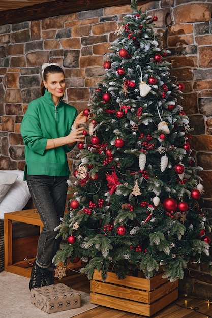 Mulher feliz de suéter verde pendura bola de brinquedo em uma árvore de Natal