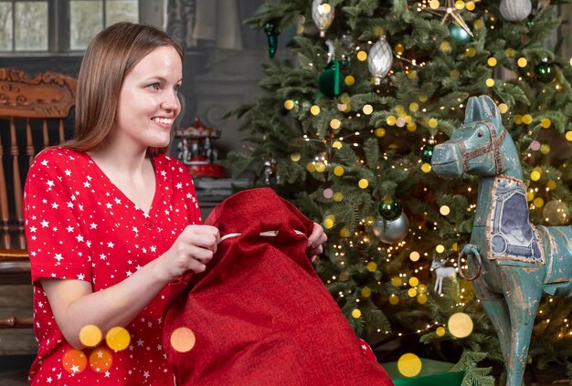Mulher feliz desempacotando o saco de natal vermelho do papai noel perto da árvore de natal