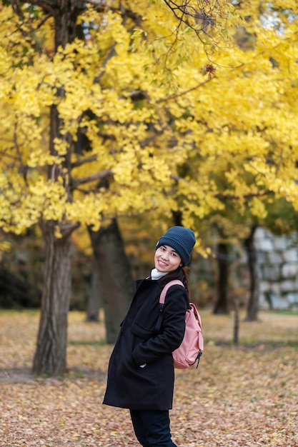 Mulher feliz desfruta no parque ao ar livre na temporada de outono viajante asiático de casaco e chapéu contra fundo de folhas amarelas de ginkgo