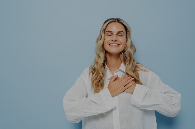 Mulher feliz e agradecida satisfeita com cabelo loiro ondulado em uma camisa branca grande demais, gesticulando apreço, amor e gratidão, segurando as mãos juntas no peito, olhos fechados, isolado sobre a parede azul