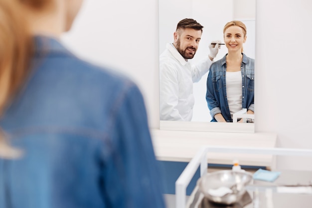 Mulher feliz e positiva olhando para seu reflexo e sorrindo ao visitar um cirurgião plástico