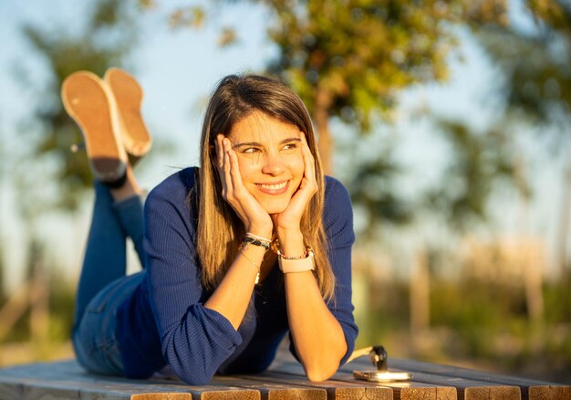 Foto mulher feliz e relaxada sorrindo enquanto está deitada em um banco ao ar livre em um parque durante o pôr do sol