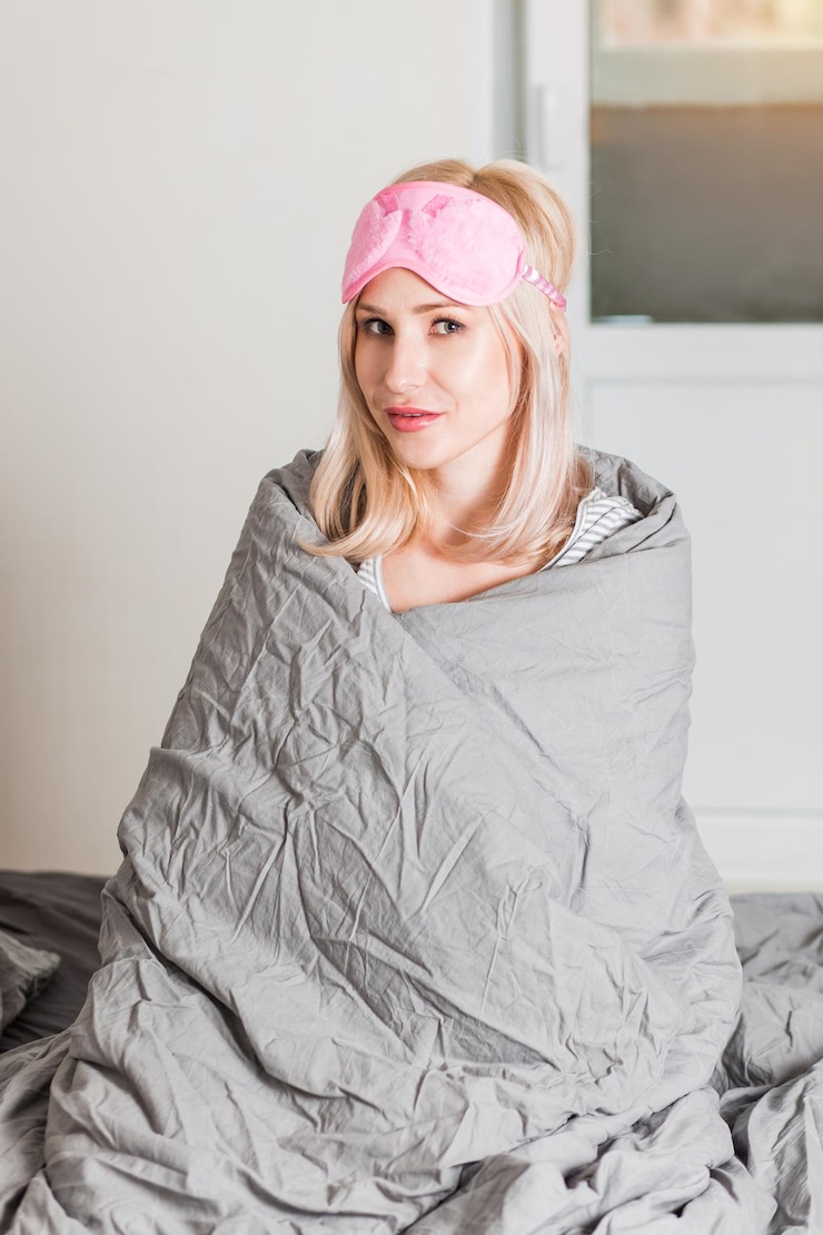 Foto mulher feliz e satisfeita com a máscara de dormir rosa na cabeça, sentando os olhos fechados na cama coberta com um cobertor cinza macio. linda mulher passar um tempo na sala. descanse, relaxe, fim de semana, conceito de bom humor.