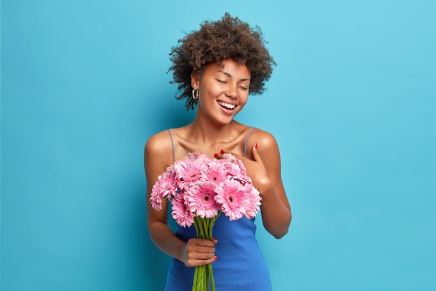 Mulher feliz e satisfeita curtindo um encontro romântico recebe buquê de flores gérbera do namorado fecha os olhos de prazer isolado sobre a parede azul fica muito grata