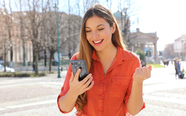 Mulher feliz e satisfeita segurando um telefone inteligente, lendo boas notícias ao ar livre e comemorando na rua da cidade
