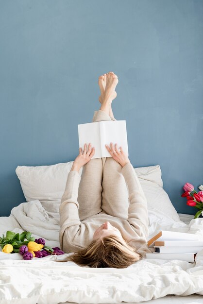 Mulher feliz e sorridente deitada na cama de pijama, com prazer, curtindo flores e lendo um livro