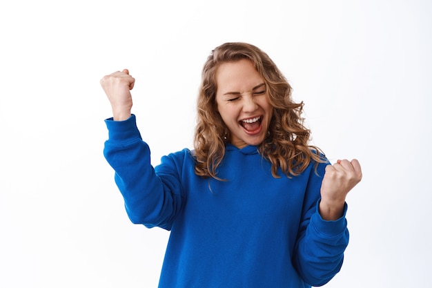 Foto mulher feliz e sortuda comemorando, faça dança do campeão, grite de alegria e satisfação após ganhar o prêmio, alcance a meta e o triunfo, de pé sobre a parede branca