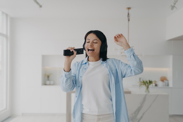 Mulher feliz em fones de ouvido cantando karaokê usando o controle remoto da TV como microfone relaxando em casa