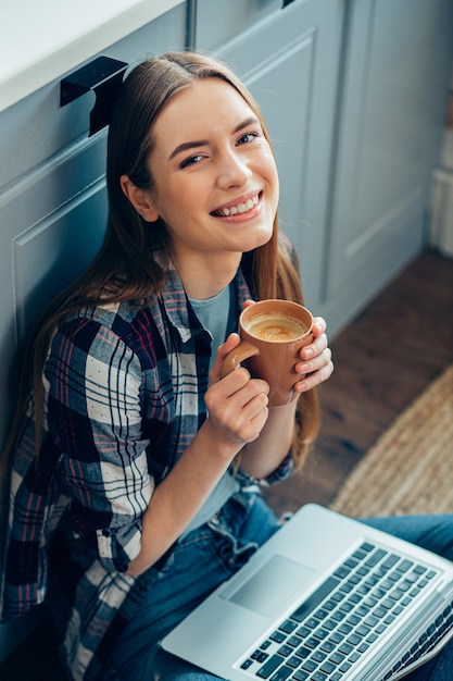 Mulher feliz em roupas casuais sentada no chão com um laptop e segurando uma caneca com café