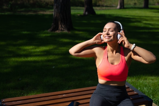 Mulher feliz em roupas esportivas com fones de ouvido, curtindo música enquanto relaxa no parque