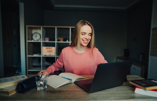 Mulher feliz estudando online em casa com laptop e livros olhando para a tela e sorrindo