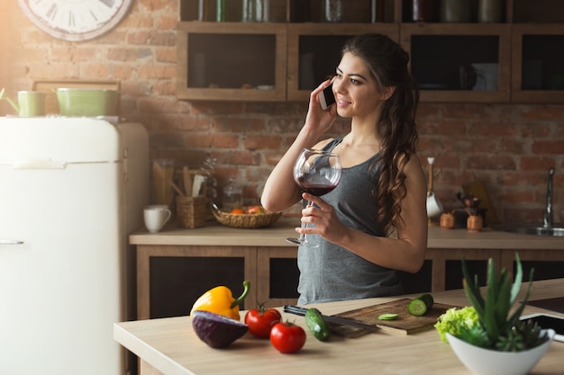 Mulher feliz falando no telefone e bebendo vinho enquanto cozinha alimentos saudáveis na cozinha do sótão em casa em dia ensolarado. Preparando salada de legumes.