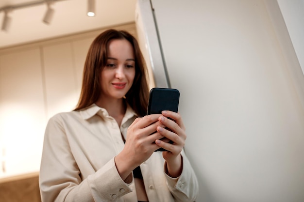 Mulher feliz freelancer em roupas casuais usando telefone em pé na cozinha de casa durante o freio de trabalho