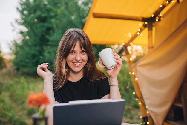 Mulher feliz freelancer usando laptop e bebendo café em uma barraca de glamping aconchegante em um dia ensolarado Barraca de acampamento de luxo para férias de verão ao ar livre e férias Conceito de estilo de vida