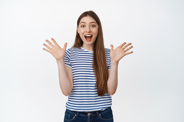 Mulher feliz gritando de alegria e surpresa, apertando as mãos e sorrindo espantado, de pé na camiseta contra fundo branco.