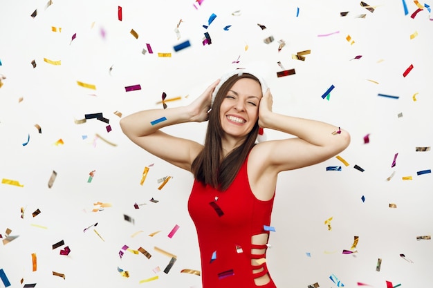 Mulher feliz jovem bonita caucasiana com sorriso encantador usando vestido vermelho e chapéu de Natal de mãos dadas na cabeça, isolada no fundo branco e confetes. Menina Santa. Conceito de feriado de ano novo 2018.