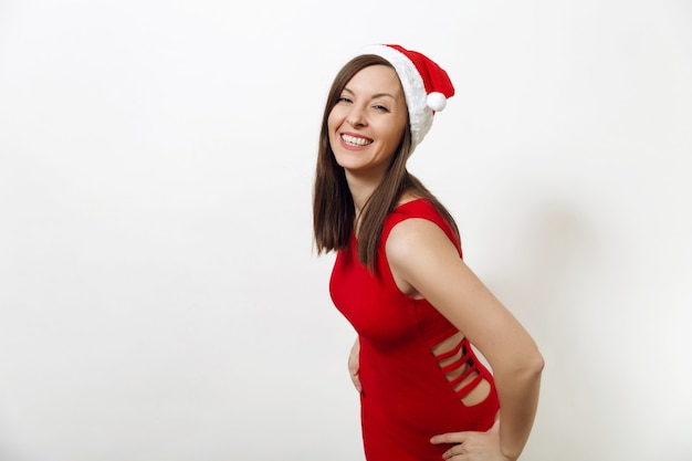 Mulher feliz jovem caucasiana bonita com sorriso encantador usando vestido vermelho e chapéu de Natal em fundo branco. Retrato isolado da menina Santa. Conceito de feriado de ano novo 2018. Olhando a câmera.