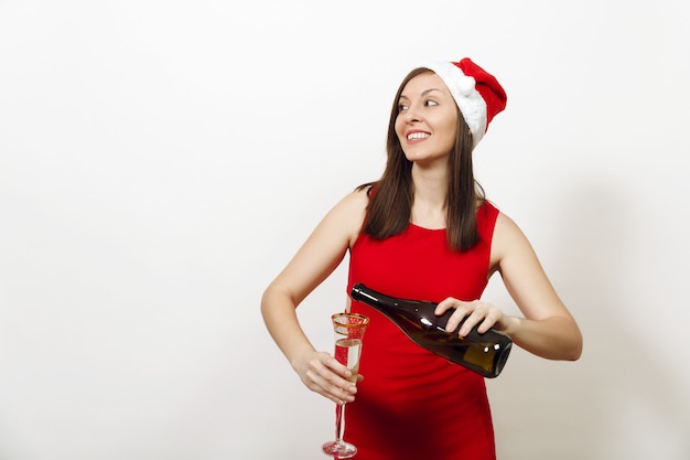 Mulher feliz jovem caucasiana bonita com um sorriso encantador no vestido vermelho e chapéu de Natal derramando champanhe da garrafa no copo no fundo branco. Santa menina isolada. Feriado de ano novo de 2018