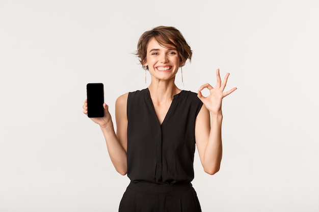 Mulher feliz jovem satisfeita mostrando a tela do celular e o gesto certo, garantir ou recomendar algo, branco de pé.