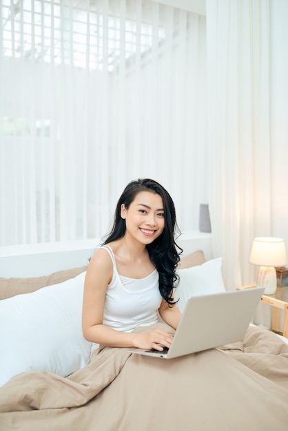 Mulher feliz lendo notícias enquanto relaxa na cama com um laptop pela manhã