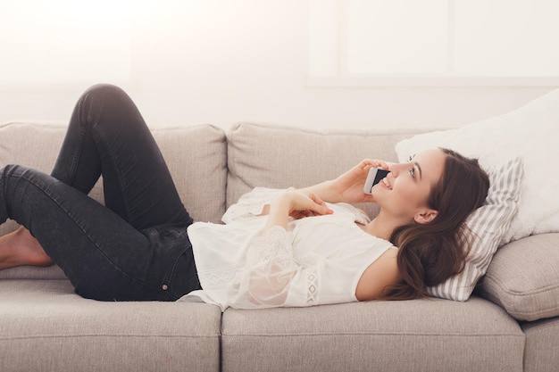 Mulher feliz ligando no celular, deitada no sofá bege em casa, closeup, copie o espaço