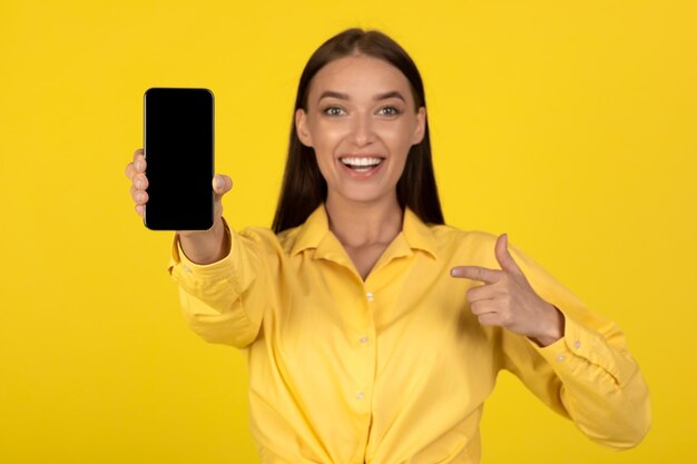 Mulher feliz mostrando a tela vazia do smartphone sobre fundo amarelo