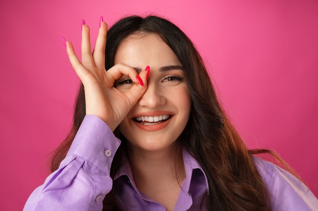 Mulher feliz mostrando um gesto de ok e olhando por entre os dedos contra um fundo rosa