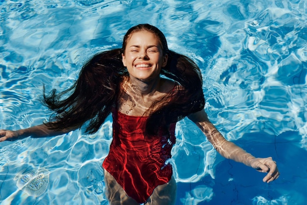 Mulher feliz nadando na piscina de fato de banho vermelho com cabelo longo solto relaxada com as mãos levantadas à luz do sol