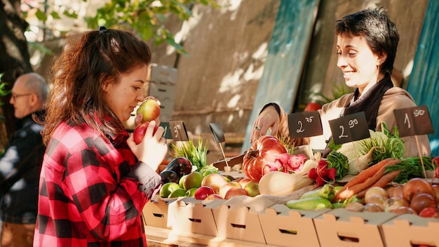 Foto mulher feliz olhando para maçãs suculentas para comprar no mercado de agricultores, verificando produtos saudáveis no estande do mercado local. cliente feminino visitando fornecedor, produtos biológicos frescos. tiro portátil.