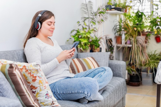 Mulher feliz ouve e assiste conteúdo do telefone sentado em um sofá em casa