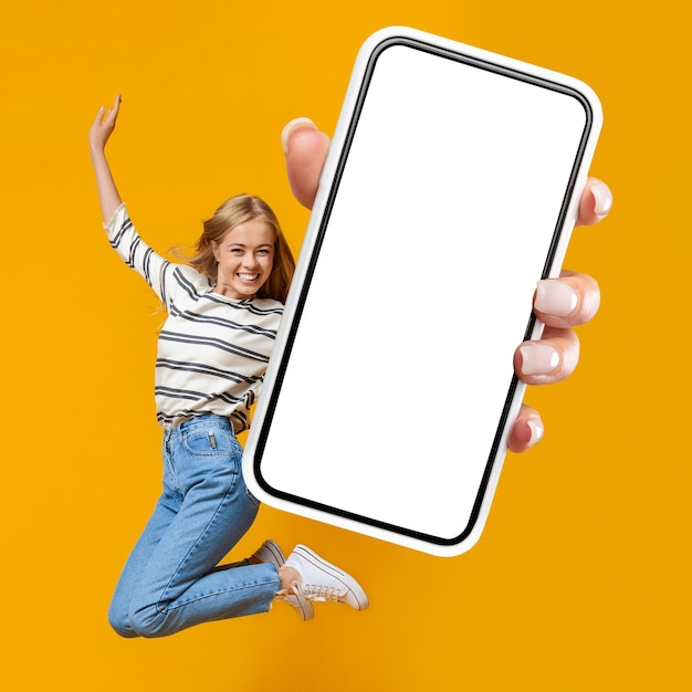 Mulher feliz pulando no ar em fundo laranja mostrando smartphone