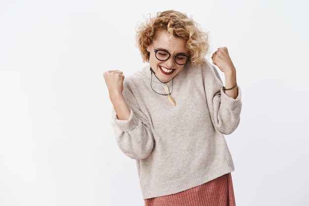 Mulher feliz, realizada e emocionada, alegre, celebrando a vitória ou o sucesso, cerrando os punhos em triunfo, olhos fechados e sorrindo amplamente realizando o sonho sobre a parede branca