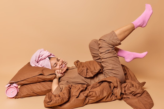 Mulher feliz relaxada mantém os olhos fechados usa pijama e toalha enrolada na cabeça se estende na cama depois de acordar gosta de bom dia roupas de cama frescas isoladas sobre fundo marrom conceito de acordar