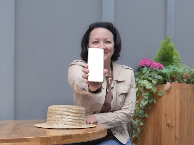 Foto mulher feliz se senta à mesa em um café e mostra a tela branca do telefone.