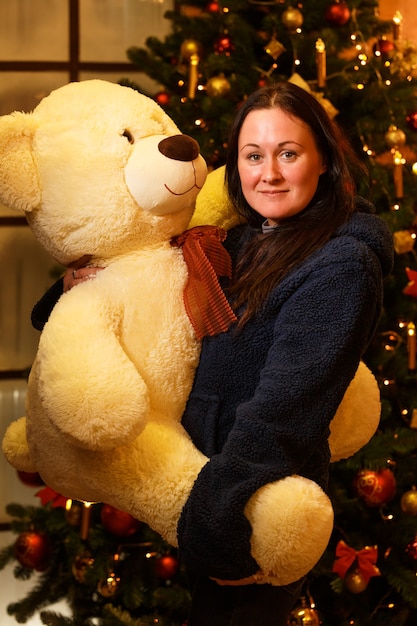 Mulher feliz segurando um grande urso de pelúcia nos braços perto da árvore de Natal