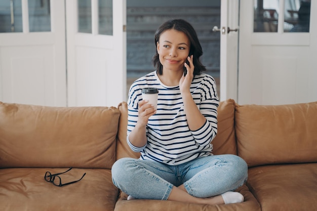 Mulher feliz segurando uma conversa de chamada de smartphone sentada no sofá com uma xícara de café na sala de estar