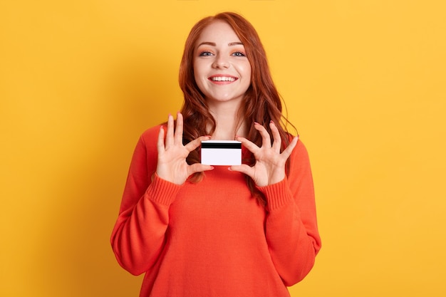 Mulher feliz segurar o cartão de crédito nas mãos, pronta para fazer compras, vestindo suéter laranja, posando isolado sobre fundo amarelo, cliente do banco, mostrando o cartão de crédito dela.