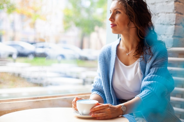 Mulher feliz sem foco com cabelo longo ondulado em uma camisola quente, segurando uma xícara de café com leite azul quente. Bebida e cor da moda. O café com leite azul é feito com flores de ervilha-borboleta. Bebida de ervas saudáveis