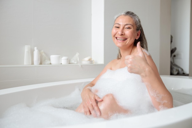 Foto mulher feliz sentada na banheira no banheiro brilhante com espuma branca, sorrindo e olhando para a câmera