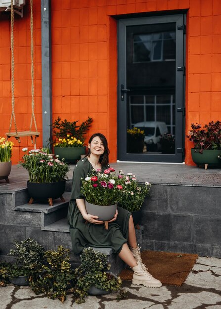 Foto mulher feliz sentada na escada de uma floricultura segurando um grande vaso de flores com flores de ranúnculo