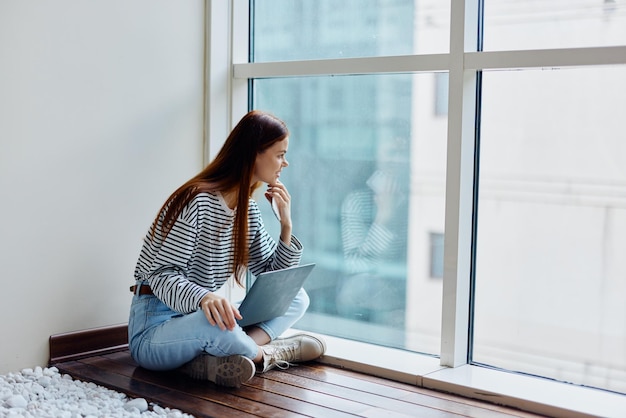 Mulher feliz sentada no chão perto das janelas com seu laptop e segurando seu telefone olhando para ele uma vista da cidade grande o conceito de trabalhar e aprender online